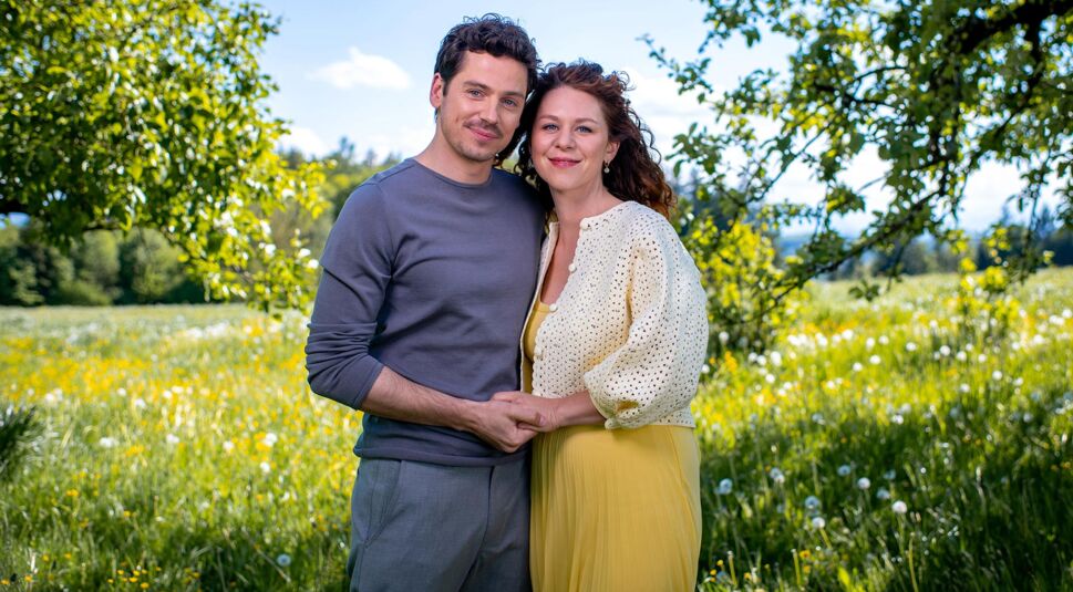 Auf dem Bild zu Sturm der Liebe Staffel 22 ist das neue Traumpaar der Staffel in einer sommerlichen Wiesenlandschaft zu sehen. Die beiden stehen eng beieinander, halten sich an den Händen und lächeln in die Kamera.