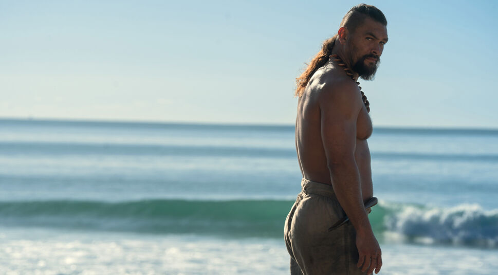 Auf dem Bild in dem Artikel zum „Chief of War“-Ende erklärt steht ein muskulöser, langhaariger Mann mit freiem Oberkörper am Strand und blickt über die Schulter. Die Szene ist bei Tageslicht aufgenommen.