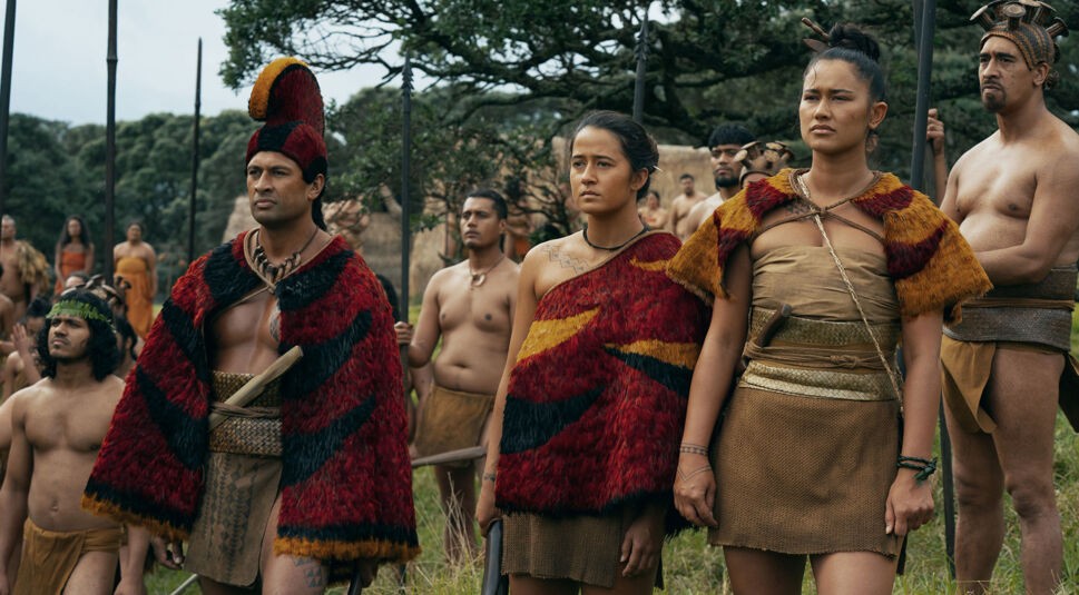 Auf dem Bild in dem Artikel zum „Chief of War“-Ende erklärt steht eine größere Gruppe Menschen in einer Wiese. Im Vordergrund sind drei Personen mit roten Umhängen zu sehen, dahinter stehen weitere mit Speeren.