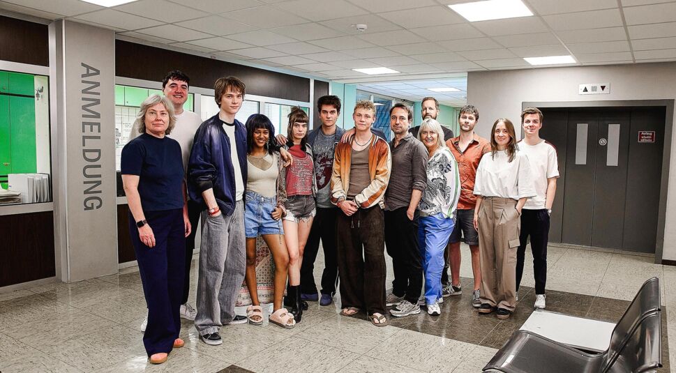 Auf dem Bild in dem Artikel zu Club der roten Bänder – Die neue Generation ist der Cast und die Crew in einem Flur zu sehen. Die Gruppe besteht aus Jugendlichen und Erwachsenen, die zusammenstehen und in die Kamera lächeln.