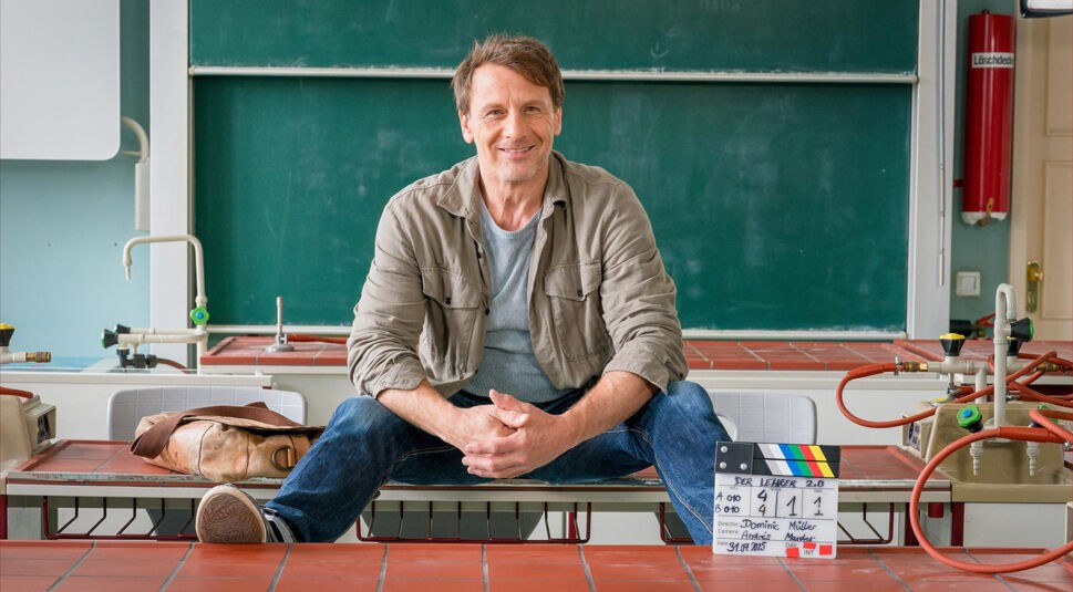 Auf dem Bild in dem Artikel zur Der Lehrer-Besetzung sitzt ein Mann auf einem Schultisch vor einer grünen Tafel. Neben ihm liegt eine Lederumhängetasche, vor ihm steht eine Filmklappe mit dem Serientitel.