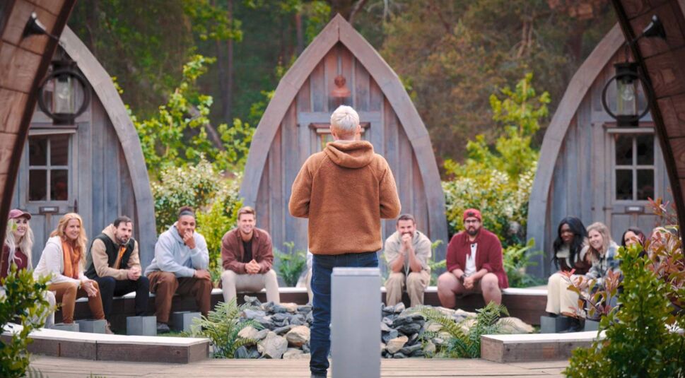 In den GigaTV-Highlights im Oktober 2025 ist eine Szene aus der Reality-Show „Werwölfe – Das Spiel von List und Täuschung“ zu sehen. Eine Gruppe von Teilnehmer:innen sitzt im Halbkreis im Außenbereich vor kleinen Holzhütten. Eine Person mit brauner Fleecejacke steht mit dem Rücken zur Kamera und spricht die Gruppe an.