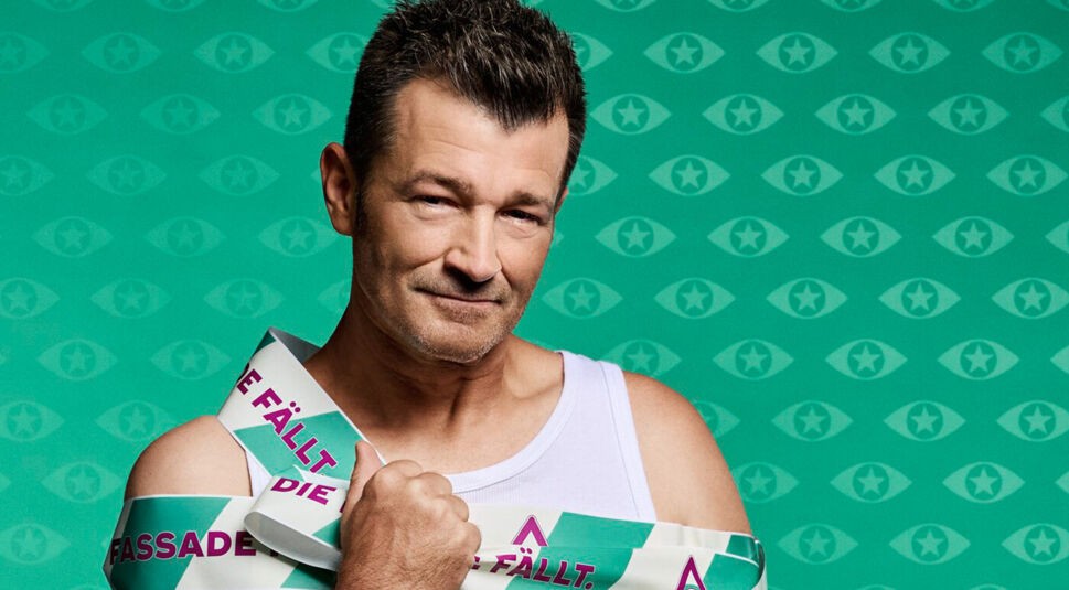 Ein männlicher Kandidat von Promi Big Brother 2025 trägt ein weißes Tanktop und blickt selbstbewusst in die Kamera. Er ist in grün-weißes Klebeband mit der Aufschrift „Die Fassade fällt“ eingewickelt.