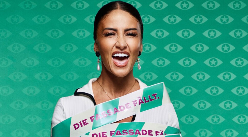 Eine Kandidatin von Promi Big Brother 2025 mit dunklen Haaren, weißen Ohrringen und weißem Outfit blickt in die Kamera. Sie ist mit grün-weißem Klebeband umwickelt, auf dem der Slogan „Die Fassade fällt“ steht.