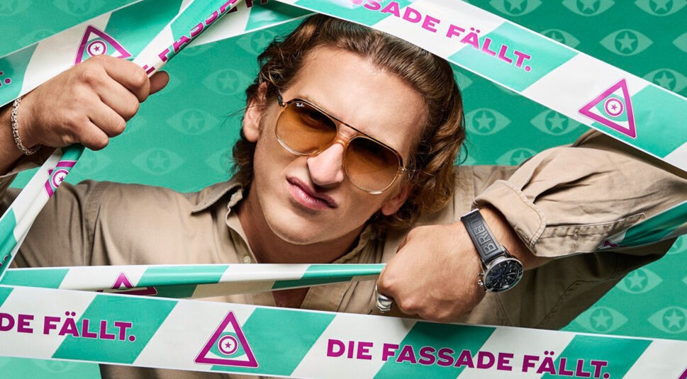 Erik, bekannt als SatansBratan, trägt auf dem Bild zu Promi Big Brother 2025 ein beiges Hemd und eine Sonnenbrille. Er schaut durch das Absperrband mit dem Schriftzug „Die Fassade fällt“.