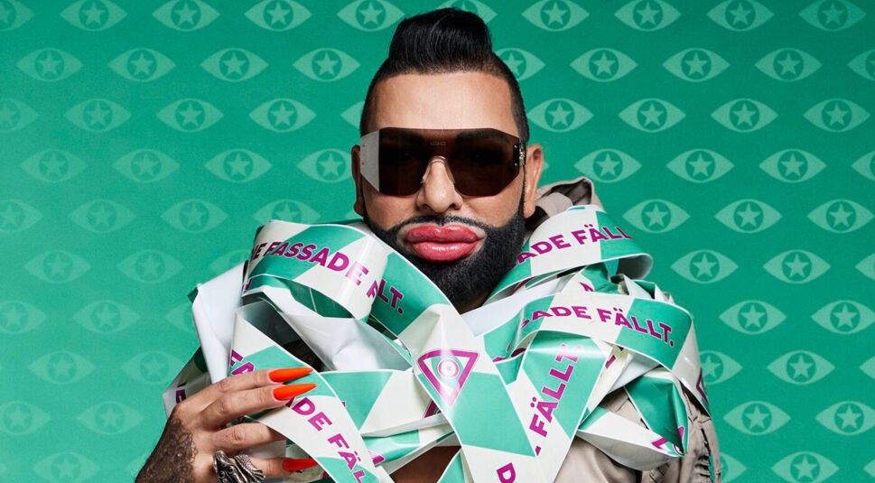 Auf dem Bild zu Promi Big Brother 2025 ist Harald Glööckler als Kandidat zu sehen. Er trägt auffällige Sonnenbrillen, extravagantes Make-up und ist in ein Band mit der Aufschrift „Die Fassade fällt“ gehüllt. Vor einem türkisfarbenen Hintergrund mit Augen- und Sternsymbolen posiert er selbstbewusst und exzentrisch in typischer Glööckler-Manier.