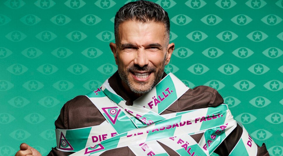Marc Terenzi posiert auf dem Bild zu Promi Big Brother 2025. Das Absperrband mit dem Slogan „Die Fassade fällt“ verläuft über sein Oberteil.