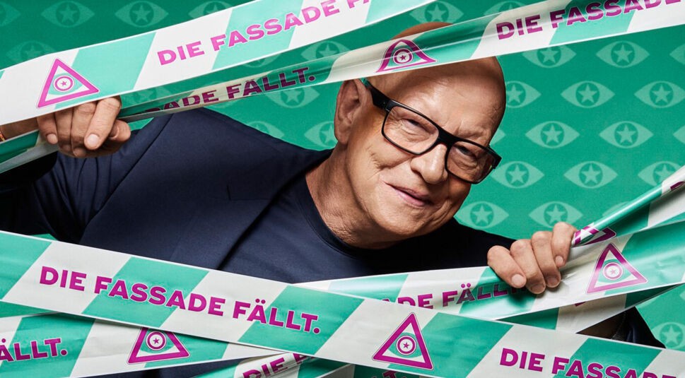 Ein männlicher Kandidat von Promi Big Brother 2025 mit schwarzer Brille und dunklem Outfit schaut aus dem Klebeband mit dem Spruch „Die Fassade fällt“ hervor.