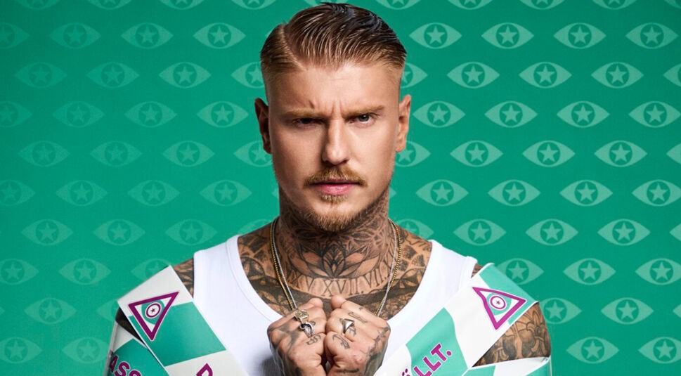 Paco Herb steht im Zentrum des Promi Big Brother 2025 Bildes. Mit verschränkten Fäusten und ernstem Blick steht er umgeben von den typischen „Die Fassade fällt“-Bändern vor dem Reality-Show-Hintergrund.