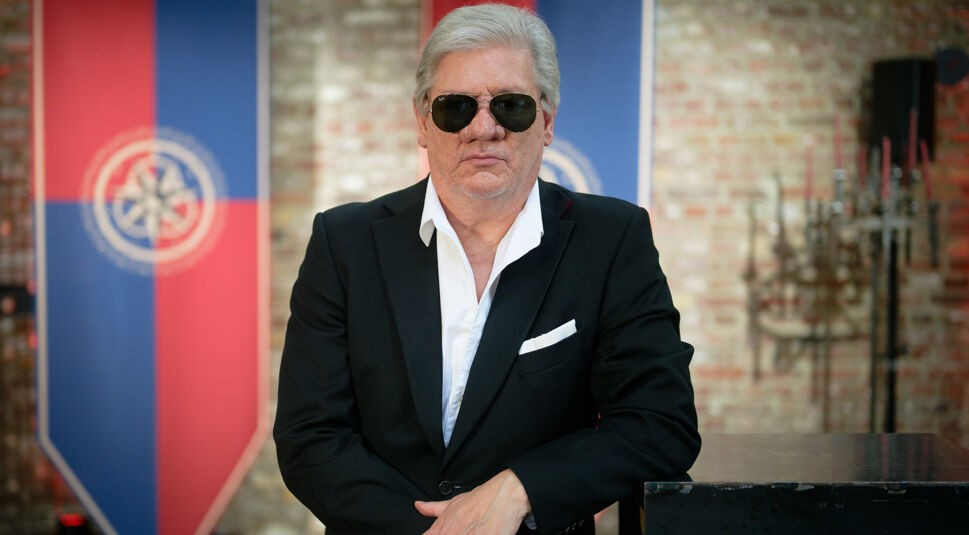 Auf dem Bild in dem Artikel zu Promis unter Palmen 2026 ist Claude-Oliver Rudolph in einem schwarzen Anzug mit weißem Hemd und Sonnenbrille zu sehen. Er steht mit verschränkten Armen vor einer rustikalen Backsteinwand.