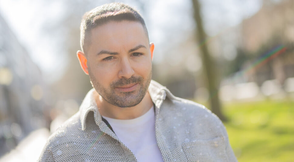 Auf dem Bild in dem Artikel zu Promis unter Palmen 2026 ist Menderes Bagci in einer silber glänzenden Jacke vor einem sonnigen, verschwommenen Stadtpark-Hintergrund zu sehen. Er lächelt leicht in die Kamera.
