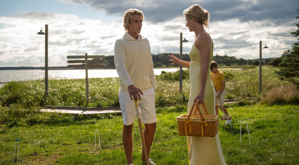 In dieser Szene aus Solange wir lügen Staffel 2 stehen Joseph Zada als Johnny Sinclair Dennis und Mamie Gummer als Carrie Sinclair auf einer sonnigen Wiese mit Blick auf einen See. Beide tragen helle, gelblich-weiße Kleidung und wirken angespannt im Gespräch. Im Hintergrund ist ein Kind beim Sammeln zu sehen. Die Atmosphäre ist ruhig, aber leicht angespannt.