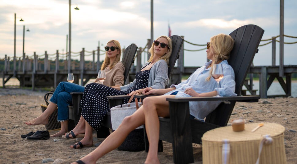 Am Strand sitzen in Solange wir lügen Caitlin FitzGerald als Penny Sinclair, Candice King als Bess Sinclair und Mamie Gummer als Carrie Sinclair entspannt in dunklen Holzstühlen. Alle drei tragen Sonnenbrillen und trinken Roséwein, während sie auf das Wasser blicken. Die Szene wirkt ruhig, stilvoll und vermittelt sommerliche Leichtigkeit vor maritimer Kulisse.