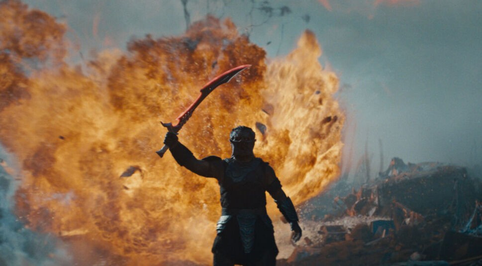 Predator: Badlands: Eine gepanzerte Kriegergestalt steht vor einer gewaltigen Explosion und hält triumphierend ein rotes, gekrümmtes Schwert in die Höhe. 