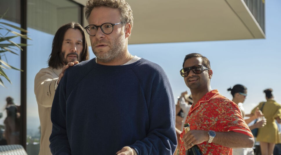 Seth Rogen, Aziz Ansari und Keanu Reeves in Good Fortune: Ein Mann in beigem Trenchcoat legt hinterrücks die Hand auf die Schulter eines verdutzt dreinblickenden Mannes mit Brille.