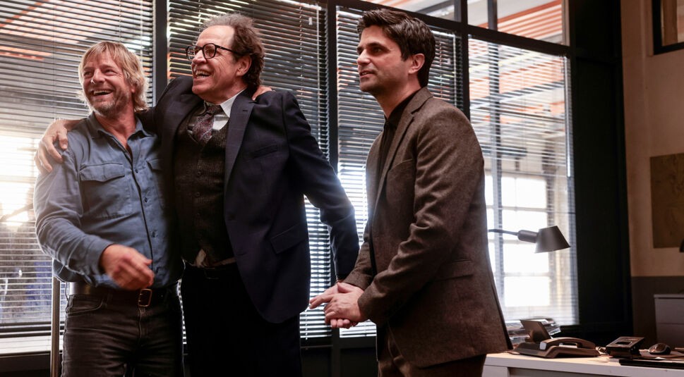 In einer Szene aus Der letzte Bulle stehen drei Männer in einem Büro. Links lacht Mick Brisgau, gespielt von Henning Baum, in einem blauen Hemd. In der Mitte ein fröhlicher Mann im Anzug, rechts ein ernsterer Kollege.