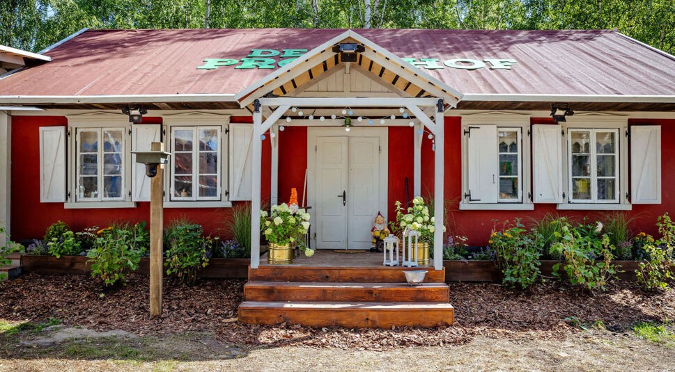 Auf diesem Bild zur Reality-Show Der Promihof ist der rot gestrichene Bauernhof mit weißem Giebeldach und dekorativem Eingangsbereich zu sehen. Vor der zentralen Doppeltür stehen Blumen, Gartenfiguren und Laternen, was eine einladende Atmosphäre schafft. Das Schild „Der Promihof“ ist groß auf dem Dach angebracht und signalisiert den zentralen Drehort der Show.