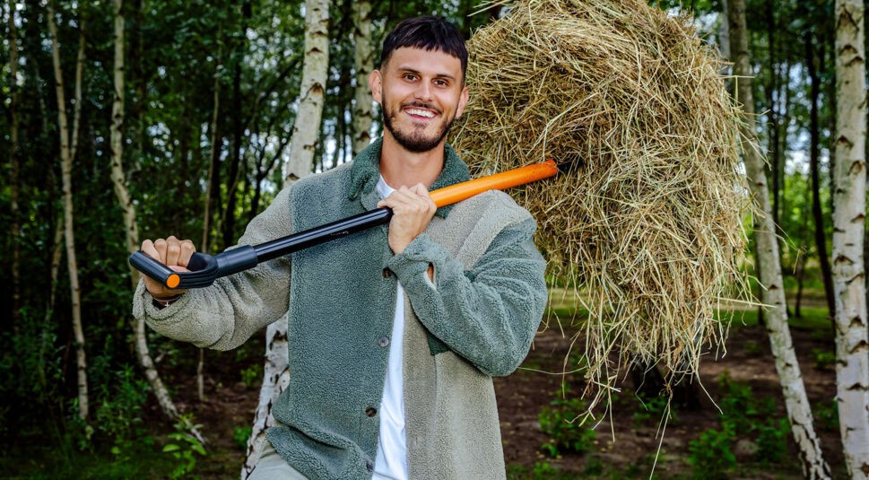 Auf dem Bild zur Sendung Der Promihof ist Nino Sifkovits zu sehen, der als Aufpasser auf dem Hof arbeitet. Er steht lachend in einem Waldstück und trägt ein Bündel Heu auf einer modernen Mistgabel über der Schulter. Seine Kleidung wirkt robust und ländlich, passend zur Umgebung. Die Stimmung ist entspannt und naturverbunden.