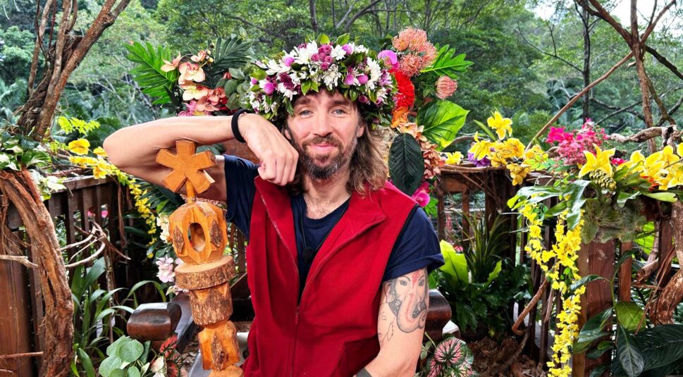 Auf dem Bild zum Dschungelcamp Gewinner 2026 ist Gil Ofarim zu sehen, der entspannt und zufrieden in einer üppigen Dschungelkulisse sitzt. Er trägt eine rote Weste, ein Blumenkranz schmückt sein Haar, in der Hand hält er das Sieger-Totem. Die Stimmung wirkt triumphierend, naturverbunden und feierlich.