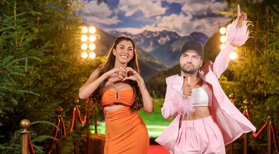 Auf dem Bild zur Realityshow Forsthaus Rampensau Germany sind Yeliz Koc und Dilara Kruse vor einer Alpen-Kulisse auf einem roten Teppich zu sehen. Yeliz trägt ein orangefarbenes Kleid und formt mit den Händen ein Herz. Daneben steht Dilara Kruse im rosa Anzug und hält eine Maske von Jimi Blue Ochsenknecht hoch. Die Stimmung wirkt fröhlich und ausgelassen.