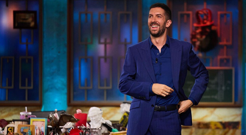 Auf dem Bild zu den internationalen Sendern und TV-Paketen bei Vodafone ist Moderator David Broncano aus der spanischen Comedy-Show „La Revuelta“ zu sehen. Er trägt einen blauen Anzug und steht lachend in einem bunt beleuchteten Studio mit dekorativem Hintergrund.