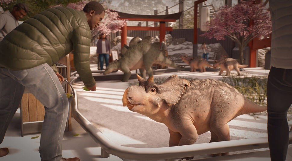 In Jurassic World Evolution 3 füttert ein Besucher ein junges Dinosaurierbaby durch ein Geländer, während im Hintergrund weitere kleine und große Dinosaurier durch ein Gehege mit Kirschbäumen laufen. Diese Szene zeigt eindrucksvoll die emotionale Tiefe und Interaktivität, die Jurassic World Evolution 3-Tipps für ein erfolgreiches Parkmanagement empfehlen.