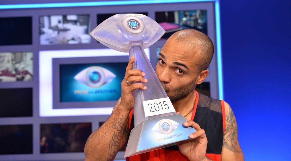 Auf dem Bild zu den Promi Big Brother Gewinner:innen ist David Odonkor zu sehen, der die Sieger-Trophäe des Jahres 2015 stolz küsst. Er trägt ein ärmelloses Sportshirt und zeigt ein zufriedenes Lächeln. Im Hintergrund sind Screens mit Livebildern aus dem Haus sowie das ikonische Promi Big Brother Logo zu erkennen.