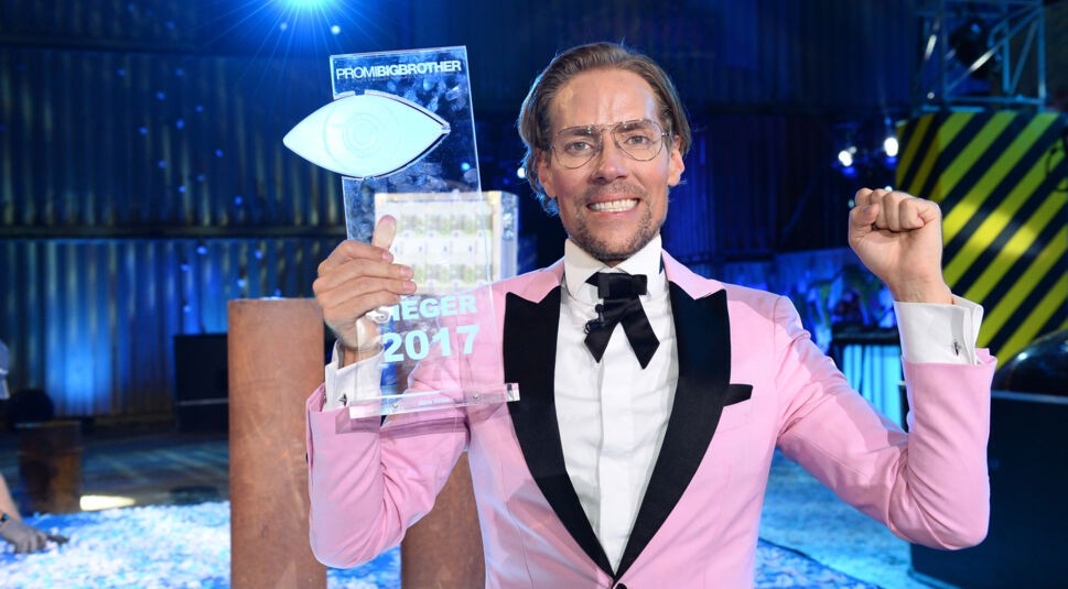 Auf dem Bild zu den Promi Big Brother Gewinner:innen ist Jens Hilbert als Sieger von 2017 zu sehen. Er trägt einen auffälligen rosa Anzug mit schwarzer Fliege und hält stolz die durchsichtige Trophäe in die Höhe. Mit erhobener Faust und strahlendem Lächeln feiert er seinen Sieg in einer bunt beleuchteten Studiokulisse.