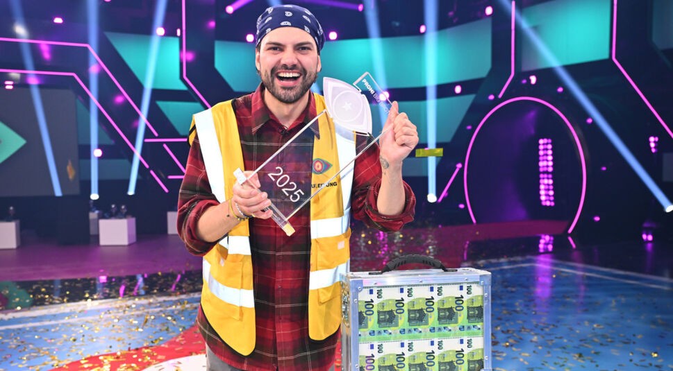 Jimi Blue Ochsenknecht strahlt als frisch gekürter Promi Big Brother Gewinner 2025 in die Kamera. In der Hand hält er stolz den durchsichtigen Siegerpokal mit der Aufschrift „2025“, neben ihm steht ein silberner Geldkoffer voller 100-Euro-Scheine. Er trägt ein kariertes Hemd, eine gelbe Sicherheitsweste und ein blaues Bandana. Die Bühne ist bunt beleuchtet und mit Konfetti bedeckt.