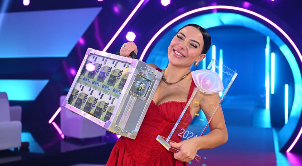 Auf dem Bild zu den Promi Big Brother Gewinner:innen ist Leyla Lahouar als Siegerin des Jahres 2024 zu sehen. Sie trägt ein auffälliges rotes Kleid und hält strahlend die gläserne Trophäe sowie einen silbernen Geldkoffer mit sichtbaren 100-Euro-Scheinen. Der futuristische Studiohintergrund ist in Pink und Blau beleuchtet, die Stimmung ist feierlich und triumphierend.