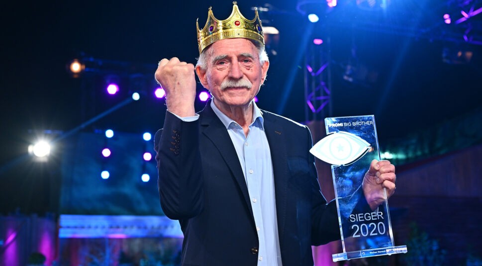 Auf dem Bild zu den Promi Big Brother Gewinner:innen ist Werner Hansch als Sieger des Jahres 2020 zu sehen. Er trägt einen dunklen Anzug mit hellblauem Hemd und eine goldene Krone auf dem Kopf. In der rechten Hand hält er die Trophäe, während er mit der linken Faust stolz in die Luft zeigt. Die Bühne ist bunt beleuchtet mit violetten und blauen Scheinwerfern.