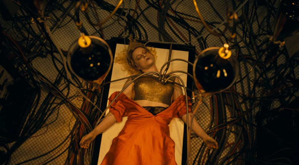 In dieser Szene aus The Bride 2026 liegt Jessie Buckley als die Braut bewusstlos auf einem Tisch, umgeben von zahlreichen Schläuchen und Kabeln. Metallene Vorrichtungen sind an ihrer Brust befestigt, während ihr rotes Kleid dramatisch inszeniert ist. Die düstere, futuristische Atmosphäre vermittelt ein Gefühl von Kontrollverlust und technischer Manipulation.