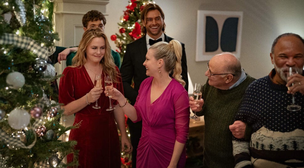 Melissa Joan Hart und Alicia Silverstone in A Merry Little Ex-Mas: Eine festlich gekleidete Gruppe stößt mit Sektgläsern vor einem geschmückten Weihnachtsbaum an; die Stimmung ist fröhlich und herzlich.