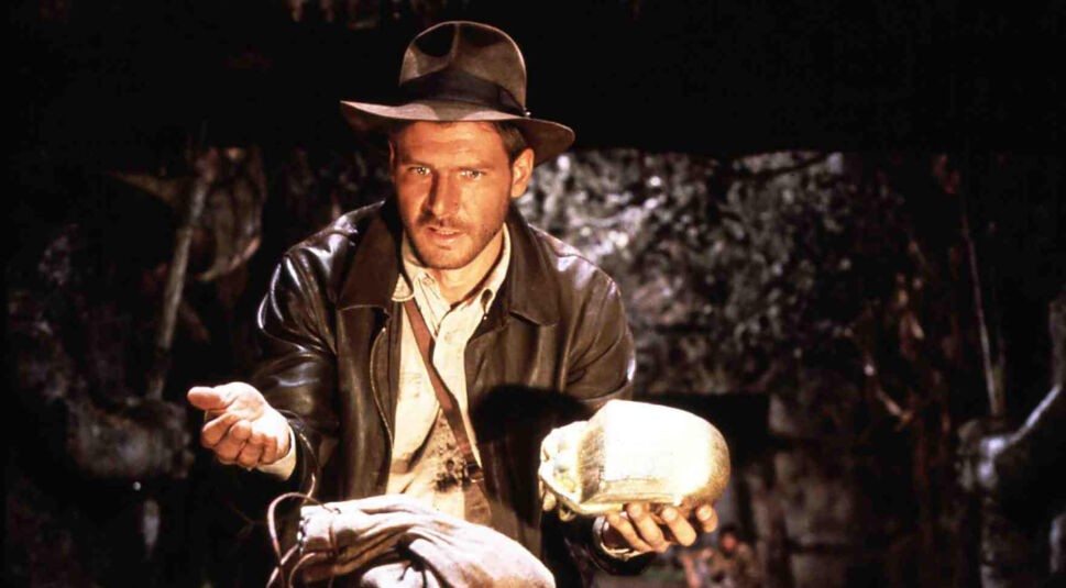 Auf dem Bild aus Jäger des verlorenen Schatzes ist Harrison Ford als Indiana Jones zu sehen – einer der legendären Actionhelden der 80er und 90er. Er trägt seinen typischen Fedora-Hut und Lederjacke, während er in einer spannungsgeladenen Szene eine goldene Statue in den Händen hält. Sein Blick ist konzentriert und entschlossen.