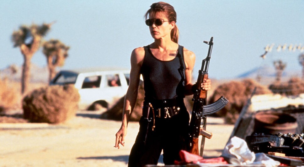 Auf dem Bild aus Terminator 2: Tag der Abrechnung ist Linda Hamilton als Sarah Connor zu sehen – bewaffnet, muskulös und entschlossen. In einem schwarzen Tanktop und mit Sonnenbrille steht sie in der Wüste, bereit zum Kampf. Sie verkörpert eine der stärksten weiblichen Actionhelden der 80er und 90er.