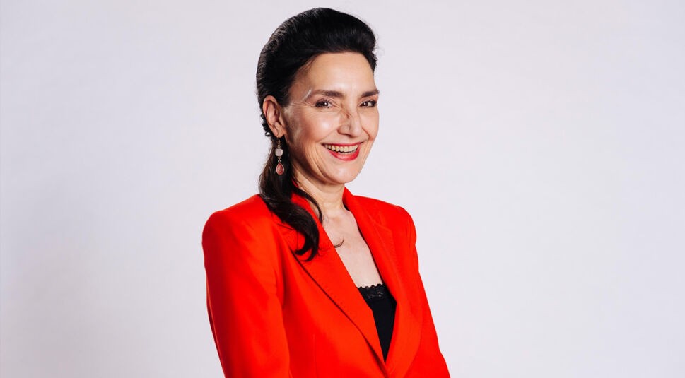 Auf diesem Bild zur Alles was zählt Besetzung ist Schauspielerin Tatjana Clasing zu sehen, die in der Serie die Rolle der Simone Steinkamp spielt. Sie trägt ein auffälliges rotes Jackett über einem schwarzen Top und lächelt herzlich in die Kamera. Ihr dunkles Haar ist elegant zurückgestylt, der Hintergrund ist hell und neutral gehalten.