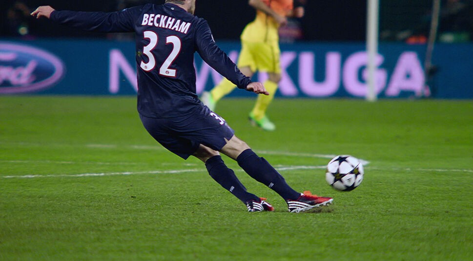 In dieser Szene aus der Fußball-Doku „Beckham“ ist Fußballlegende David Beckham während eines Spiels in Aktion zu sehen. Er trägt ein dunkelblaues Trikot mit der Rückennummer 32 und seinem Namen, während er mit voller Dynamik den Ball spielt.