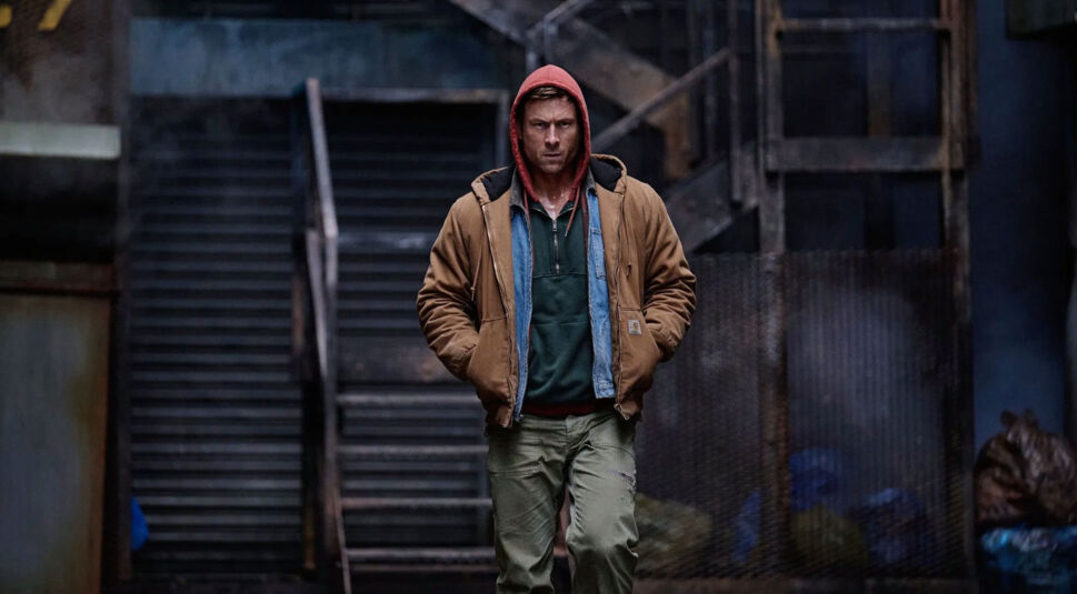 Auf dem Bild in den GigaTV-Highlights im Dezember 2025 siehst du eine Szene aus „The Running Man“: Ben Richards (Glen Powell) geht entschlossen durch eine düstere Gasse mit industriellem Hintergrund, bekleidet mit Kapuzenpulli und Jacke.