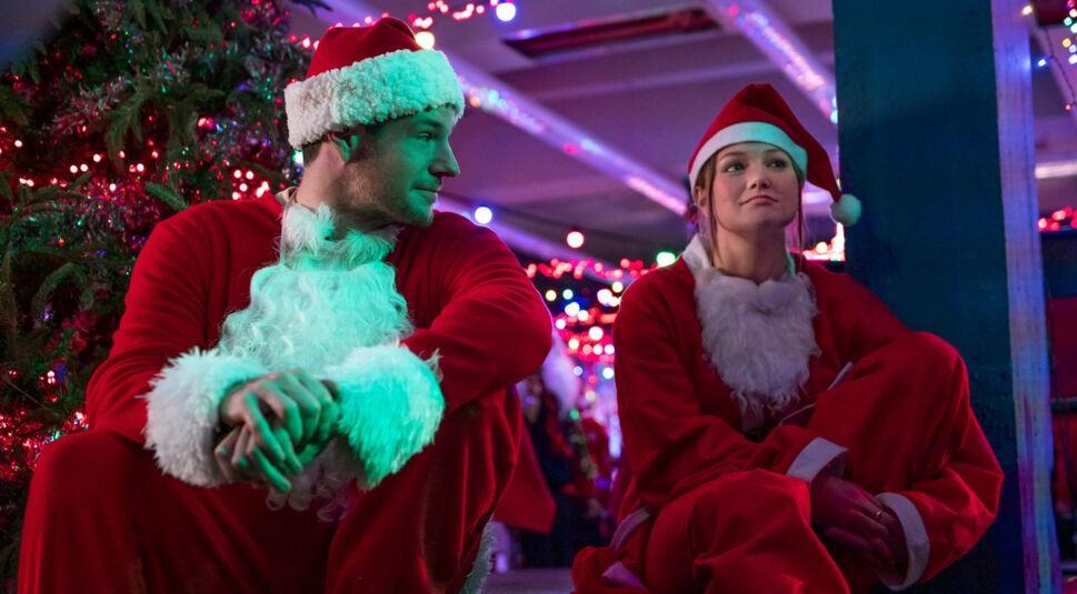 Im Bild aus Jingle Bell Heist sitzen Olivia Holt als Sophia und Connor Swindells als Nick nebeneinander in Weihnachtsmannkostümen. Die beiden wirken erschöpft, aber verbunden, während sie sich inmitten von bunten Lichterketten und einem geschmückten Weihnachtsbaum anschauen. Die Szene aus dem Netflix-Film Jingle Bell Heist kombiniert festliche Atmosphäre mit emotionaler Tiefe.