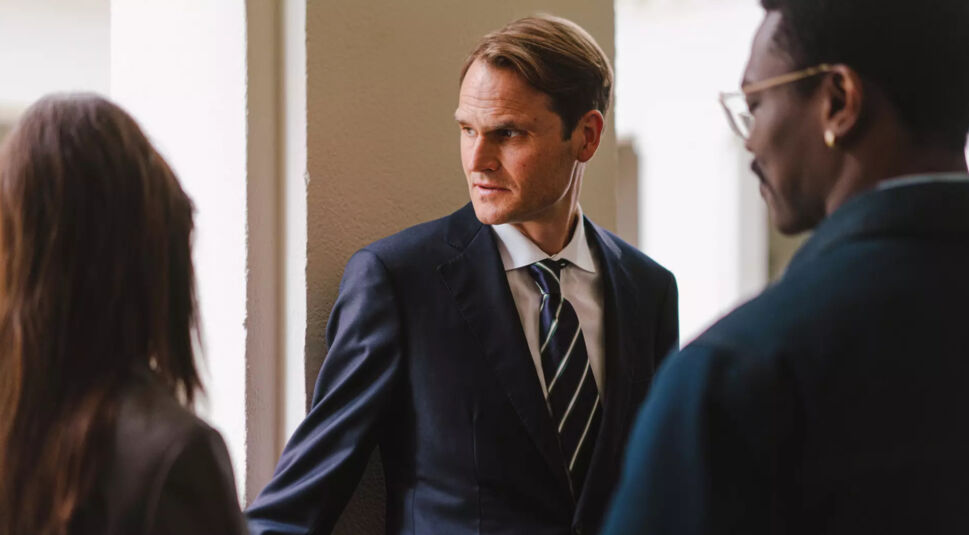 Fabian Hinrichs in Asbest: Eine Frau und zwei Männer im Gespräch in einem hellen Flur. Ein Mann im Anzug blickt ernst zur Seite.