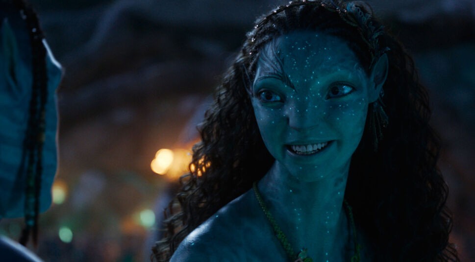 In dieser Szene aus Avatar: Fire and Ash ist die Na'vi-Figur Ronal zu sehen, gespielt von Kate Winslet. Sie lächelt freundlich und wendet sich einer Person gegenüber, die nur teilweise im Bild zu erkennen ist. Ihre Haut leuchtet blau im Licht der Umgebung. Der Alttext dient als Teil einer Avatar: Fire and Ash-Kritik.