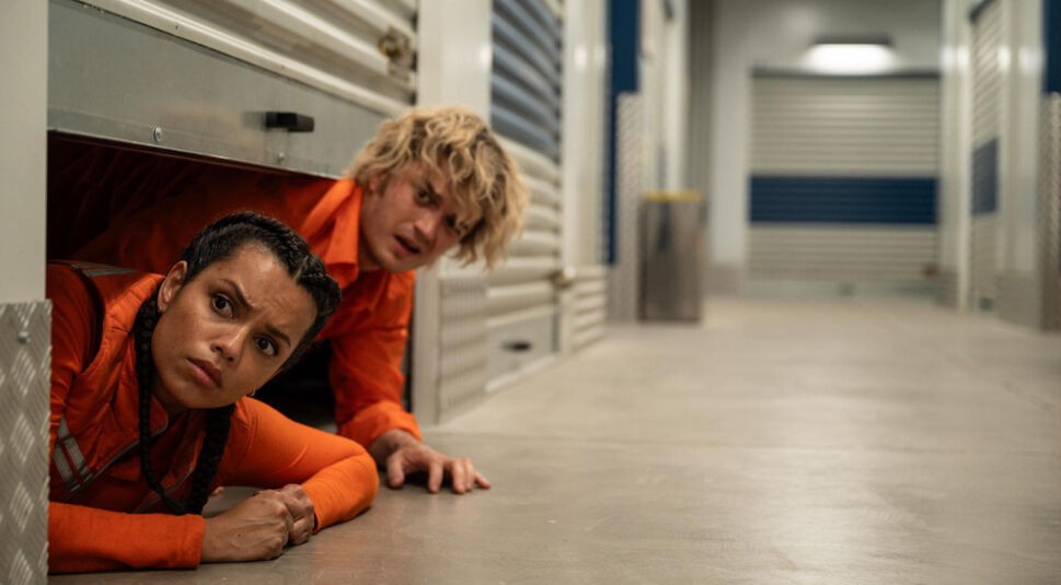 In dieser Szene aus Cold Storage kriechen Teacake, gespielt von Joey Keery, und Naomi, dargestellt von Georgina Campbell, aus einer geöffneten Lagereinheit in einen langen, sterilen Lagerhausgang. Beide tragen orangefarbene Overalls und blicken angespannt nach vorn, als würden sie sich vor etwas verstecken oder eine Gefahr beobachten. Die Stimmung ist angespannt und geheimnisvoll.