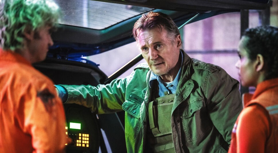 In dieser Szene aus dem Film Cold Storage steht Robert, gespielt von Liam Neeson, in schusssicherer Weste vor einem geöffneten Fahrzeugheck und spricht ernst mit Teacake (Joey Keery) und Naomi (Georgina Campbell), die orangefarbene Overalls tragen. Die Beleuchtung ist kühl und dramatisch, was die Spannung der Situation unterstreicht.