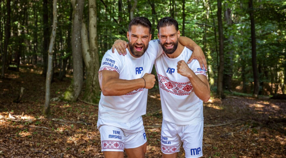 Auf diesem Bild aus der Couple Challenge 2026 stehen Aleks und Mark im Wald, beide tragen weiße Sportkleidung mit folkloristischem Muster und den Aufdruck „Team Maskulin“. Sie posieren lachend und energiegeladen mit geballten Fäusten, der Arm jeweils um die Schulter des anderen gelegt. Die Stimmung ist kämpferisch.