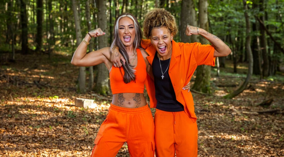 Auf dem Bild aus der Couple Challenge 2026 posieren Carina und Chika selbstbewusst im Wald. Beide tragen auffällige orangefarbene Outfits und zeigen lachend ihre angespannten Oberarme in einer Power-Pose. Ihre energiegeladene Ausstrahlung und der enge Zusammenhalt spiegeln Teamgeist und Kampfbereitschaft wider.