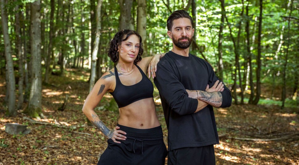 Auf dem Bild aus der Couple Challenge 2026 stehen Liza und Sebi selbstbewusst im Wald. Beide tragen schwarze Sportkleidung – Liza ein Sport-Bralette und Sebi ein langärmliges Shirt. Liza legt locker den Arm auf Sebis Schulter, während er mit verschränkten Armen in die Kamera blickt. Ihre Ausstrahlung wirkt ruhig, fokussiert und entschlossen.