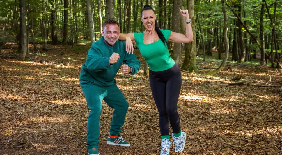 Auf dem Bild aus der Couple Challenge 2026 sind Patti und Patrick in sportlicher Siegerpose im Wald zu sehen. Patrick trägt einen grünen Fleece-Trainingsanzug, Patti ein grünes T-Shirt mit schwarzer Leggings. Beide lachen breit, zeigen starke Posen mit geballten Fäusten und strahlen pure Motivation und Wettkampfgeist aus.