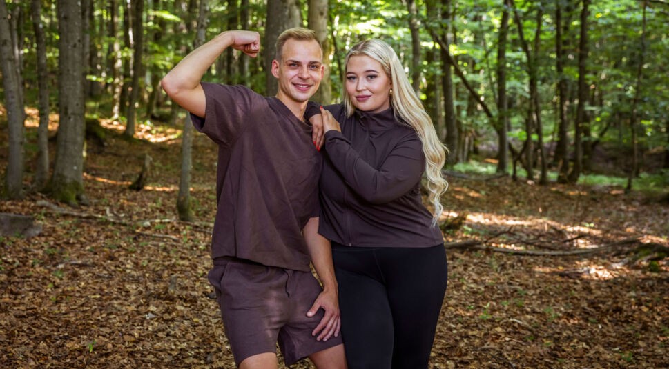 Auf dem Bild aus der Couple Challenge 2026 stehen Sophie und Hendrik in brauner Sportkleidung im Wald. Hendrik zeigt mit einem Lächeln seinen Bizeps, während Sophie sich entspannt an ihn anlehnt und selbstbewusst in die Kamera blickt. Beide wirken harmonisch und bereit für die Herausforderungen des Wettbewerbs.