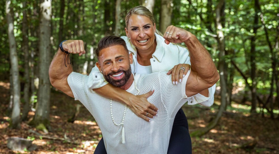 Auf dem Bild aus der Couple Challenge 2026 trägt Andrew ein weißes Netzshirt und hebt Virginia auf den Rücken, die eine weiße Jacke trägt. Beide zeigen lachend ihre Muskeln und blicken kraftvoll in die Kamera. Ihre Pose strahlt pure Energie, Teamgeist und Selbstvertrauen inmitten der Waldkulisse aus.