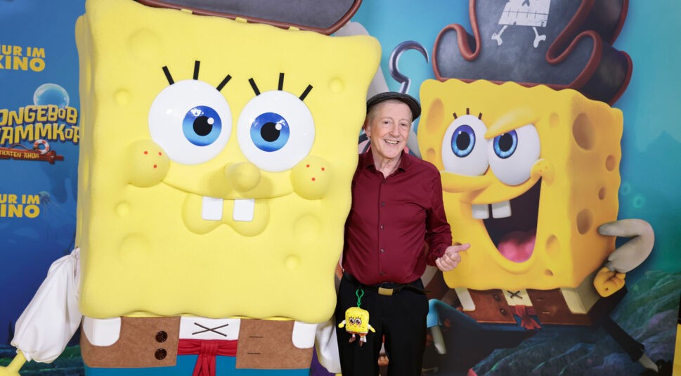 Auf dem Bild steht Santiago Ziesmer, der bekannte SpongeBob-Synchronsprecher, lächelnd neben einer lebensgroßen SpongeBob-Figur in Piratenkleidung. Er trägt ein weinrotes Hemd, eine Schiebermütze und hat ein kleines SpongeBob-Plüschtier am Gürtel. Im Hintergrund ist ein farbenfrohes Filmplakat von „SpongeBob Schwammkopf“ zu sehen. Die Stimmung ist heiter und verspielt.